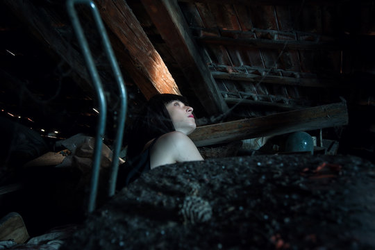 Portrait of a young brunette girl with red lipstick in an old attic, looking up to the sun ray.