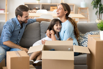 Happy family home owners on moving day concept, smiling mom hugs excited kid daughter unpacking boxes on sofa with dad in new flat, parents with child embracing on couch laughing enjoying relocation
