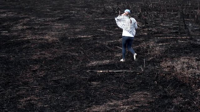 Earth after a fire/Natural disaster. A woman with a bottle of water walks along the land burned by fire. There are dead trees, burned grass around her. It's an ecological catastrophe