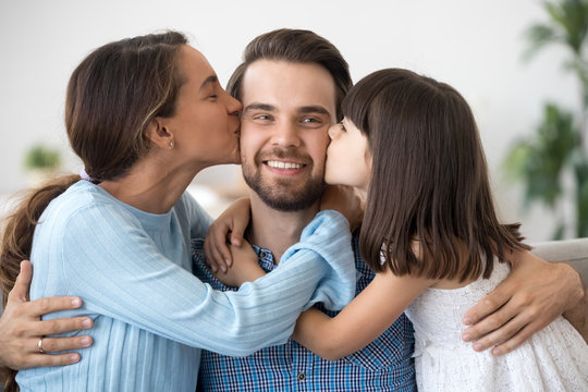 Cute kid daughter and loving wife embracing kissing young happy man on cheeks, caring beloved woman and little child girl congratulating dad husband with fathers day or birthday hugging smiling daddy