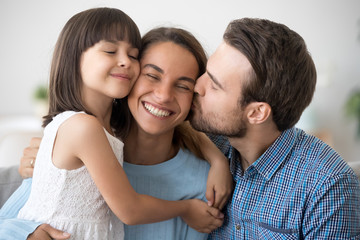 Loving husband and cute kid daughter embracing kissing happy mom wife on cheek congratulating with...