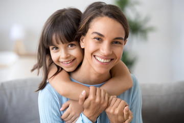 Beautiful family of single young mom and cute preschool kid daughter embracing mommy looking at camera, happy loving mother piggybacking smiling little child girl hugging mum headshot portrait