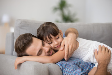 Happy father hugging cute little girl lying together resting on comfortable sofa, smiling dad...