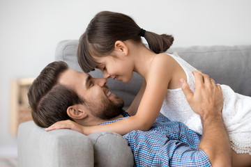 Cute little girl tenderly touching noses with happy dad lying on sofa, kid daughter and father...