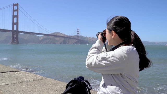 travel backpack photographer sitting on rock by sea taking photo of golden gate bridge with blue sky. young girl shooting beautiful view and zooming up the picture in camera. female relaxing by ocean
