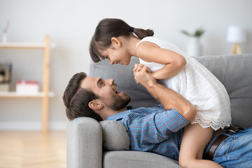 Happy dad and playful cute kid girl holding hands playing on sofa at home, father and little...