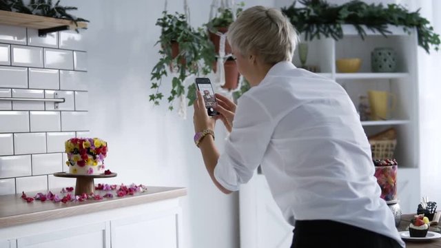Confectionery blog. Woman in white shirt photographing cake decorated with flowers. Confectioner decorated the cake ke and then takes pictures on the smartphone