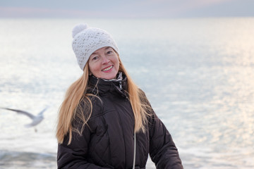 Smiling mature woman, with beautiful long hair, wearing a white knitted cap, at the beach  enjoys a healthy active lifestyle in winter time. Winter mood. Copy space.