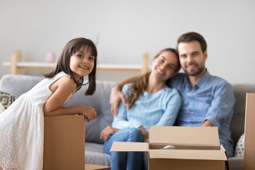 Portrait of happy kid girl posing with cardboard boxes on moving day with smiling parents at background, cute little child daughter looking at camera in new home enjoying family relocation concept
