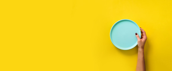 Female hand holding empty blue plate on yellow background with copy space. Healthy eating, dieting concept. Banner