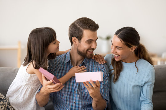 Cute Little Daughter And Loving Wife Making Funny Pleasant Surprise Congratulating Happy Husband Daddy Opening Gift Box, Excited Dad Holding Present From Family Celebrating Birthday Or Fathers Day