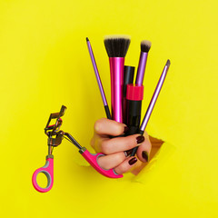 Woman hand with professional cosmetic tools for make up: brushes, mascara, lipstick, eyelash curler on yellow background. Beauty concept for cosmetics sale. Square crop