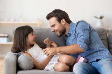 Happy daddy and playful cute kid daughter play together tickling on sofa at home, smiling dad...