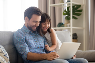 Happy father and little daughter laughing watching funny video using laptop, cheerful dad and child girl having fun playing online game on sofa together, daddy with kid relaxing at home with computer