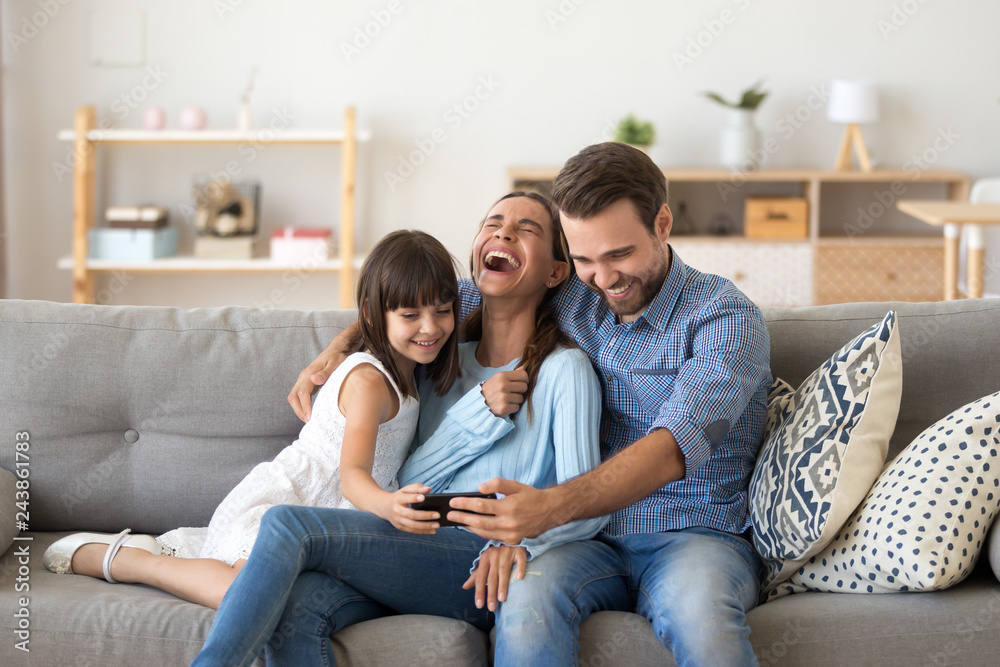 Wall mural Cheerful family vloggers with child daughter taking selfie, recording vlog, making video call on phone having fun together in living room, parents and kid girl laugh using cellphone funny online app