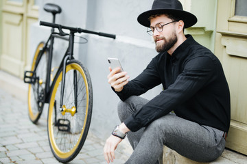 Fototapeta na wymiar Chilling after good ride. Handsome young bearded man holding mobile phone while standing near his bicycle on the street