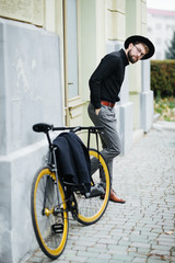 Confident and stylish. Full length of confident young bearded man adjusting eyewear and hat and looking at camera while standing near his bicycle outdoors