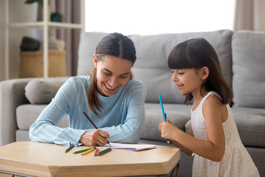Smiling mother or baby sitter helping preschool girl teaching cute kid to draw with colored pencils at home, happy child having fun drawing on paper, creative family mom and daughter hobbies concept