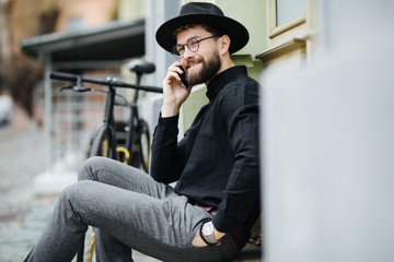Inviting friend for a ride. Happy young bearded man talking on the mobile phone and smiling while sitting near his bicycle outdoors in the street