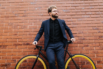 Handsome young bearded man in suit and glasses standing near his bicycle against the brick wall outdoors