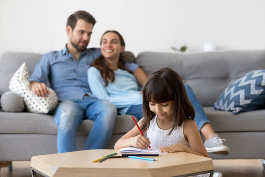 Cute smiling kid girl enjoys creative activity while parents relax at home, happy little child daughter drawing with colored pencils at table playing having fun developing imagination concentration