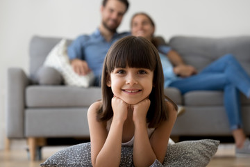 Adorable little girl looking at camera lying on warm floor relaxing with parents at home, smiling...