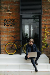 Young handsome bearded man sitting on the street stairs working on laptop near his bicycle