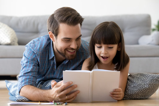 Happy caring father and surprised kid reading book together excited by interesting story, cute child girl listening to dad telling fairy tale lying on warm floor playing having fun at home together