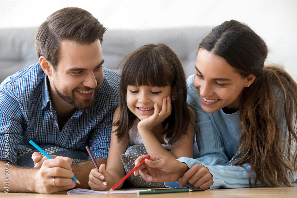 Canvas Prints Smiling caring parents and cute little daughter drawing with pencil coloring picture lying on floor, happy friendly family with kid girl play together at home, creative mom dad child weekend activity