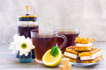 Black tea ceremony - waffles, glass full of tea, glass pot, sugar, yellow lemon, flower, tea leaves, spices on a wooden boards background