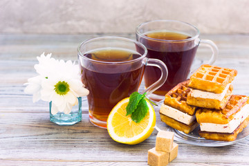Black tea ceremony - waffles, glass full of tea, glass pot, sugar, yellow lemon, flower, tea leaves, spices on a wooden boards background