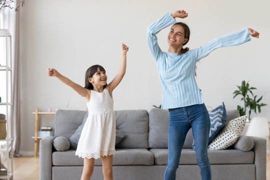 Happy Fit Sporty Mother Or Baby Sitter And Funny Active Child Daughter Having Fun Dancing Together At Home, Smiling Mom And Kid Girl Enjoy Moving To Music Raising Hands Standing In Living Room