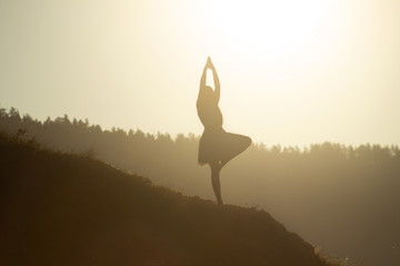 A silhouette of a young woman standing in vrikshasana, a tree pose of yoga, on sunrise