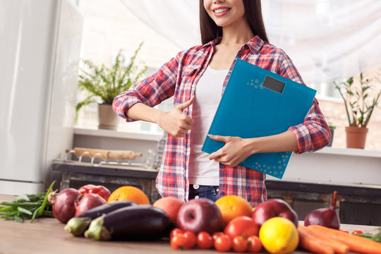 Young girl at kitchen healthy lifestyle standing near table holding scales cheerful showing thub up close-up