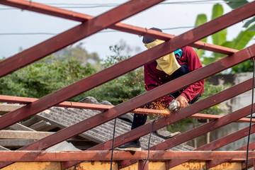 The man wearing brown shirt is cutting or sawing the metal on the roof construction by the metal saw on the top of the house in the sunny day. 