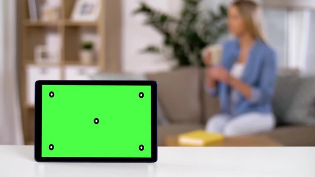 technology and objects concept - tablet computer with chroma key green screen on table and woman drinking tea on background at home
