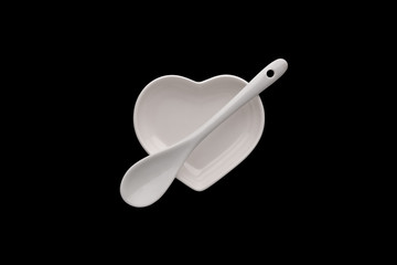 An isolated white ceramic saucer in the shape of a heart and a spoon across the plate on a black background. Modern glossy dishes. Copy space. Minimalism. Object for Valentine's Day or romantic menu.