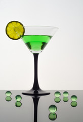 Martini glass with a green drink and a slice of lime, standing on a dark reflective table, next to the green transparent balls