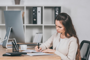 concentrated young businesswoman sitting at workplace and writing in notebook