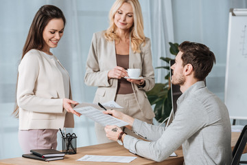 smiling mature businesswoman holding cup of coffee and looking at young colleagues working with papers in office