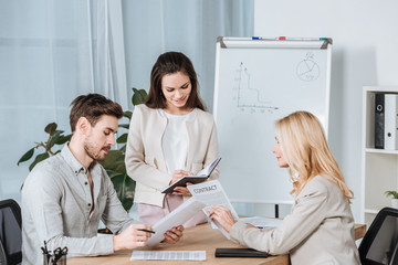 Fototapeta premium smiling young businesswoman writing in notebook and colleagues working with papers in office