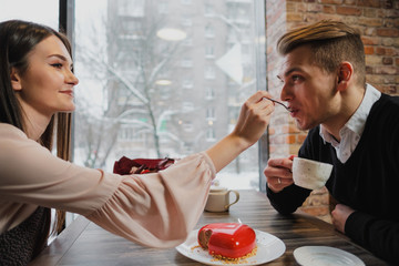 A pair of lovers, a man and a woman, eat with spoons a red heart-shaped dessert in a cafe.