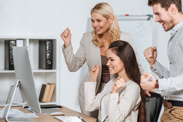 happy business mentor with smiling young colleagues shaking fists and looking at desktop computer in office