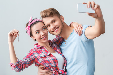 Young couple moving to new place standing hugging holding key taking selfie photos with smartphone joyful