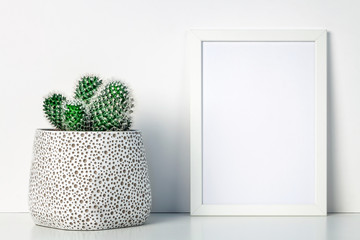 White shelf in the house with a cactus in a pot with a dot pattern and a white frame mockup. Hipster decoration. Scandinavian style. A place for your text or graphics.