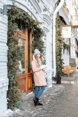 Outdoors lifestyle fashion portrait of pretty blonde girl walking on the holiday city. Smiling, drinking coffee. Wearing stylish pink coat and wide scarf. Christmas mood