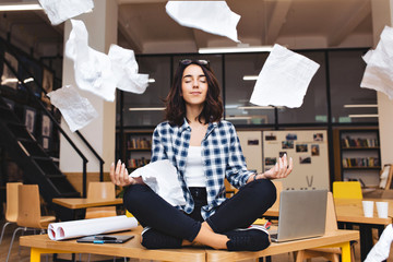 Young pretty joyful brunette woman meditating on table surround work stuff and flying papers....