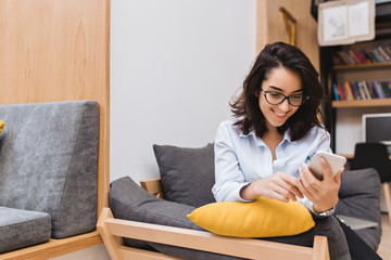 Portrait young  joyful brunette woman in black glasses chilling on couch in modern apartment. Using...
