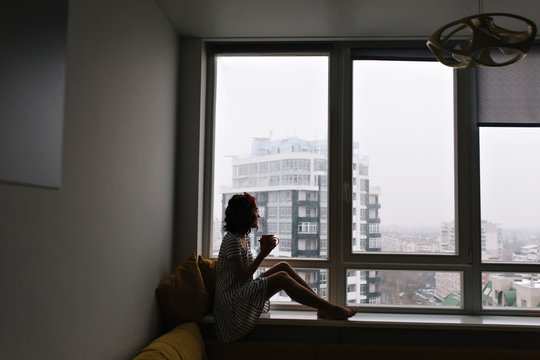 Stylish cozy image of romantic, a little bit sad pretty young woman enjoying raining weather outdoor from the window in modern apartment. Cup of tea, comfort at home