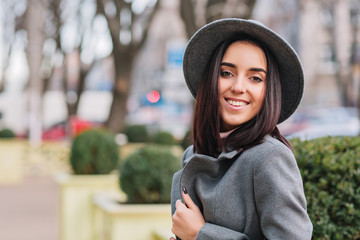 Closeup Portrait fashionable young  woman in grey hat, coat walking on street in city park. Brunette hair, smiling to camera, cheerful mood, elegant outlook. Place for text